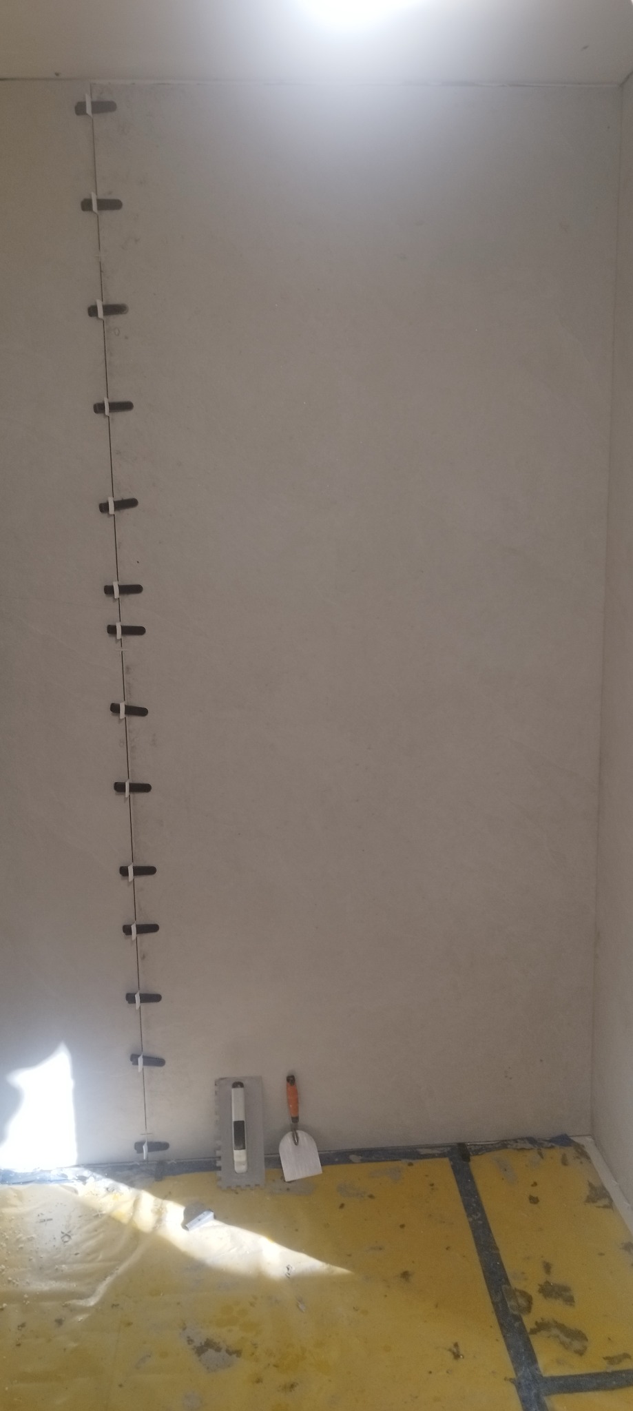 Ściana w trakcie układania płytek, widoczne krzyżyki dystansowe, narzędzia glazurnicze i zabezpieczona folią podłoga.