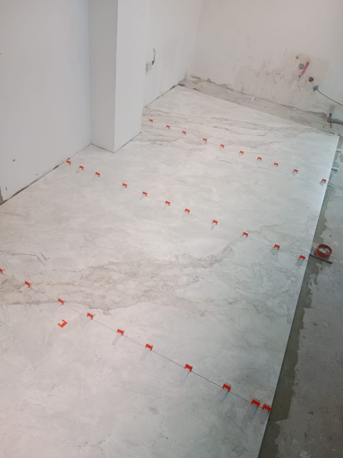 Układanie jasnych, marmurkowych płytek podłogowych z użyciem poziomujących klipsów i klinów w trakcie prac wykończeniowych.