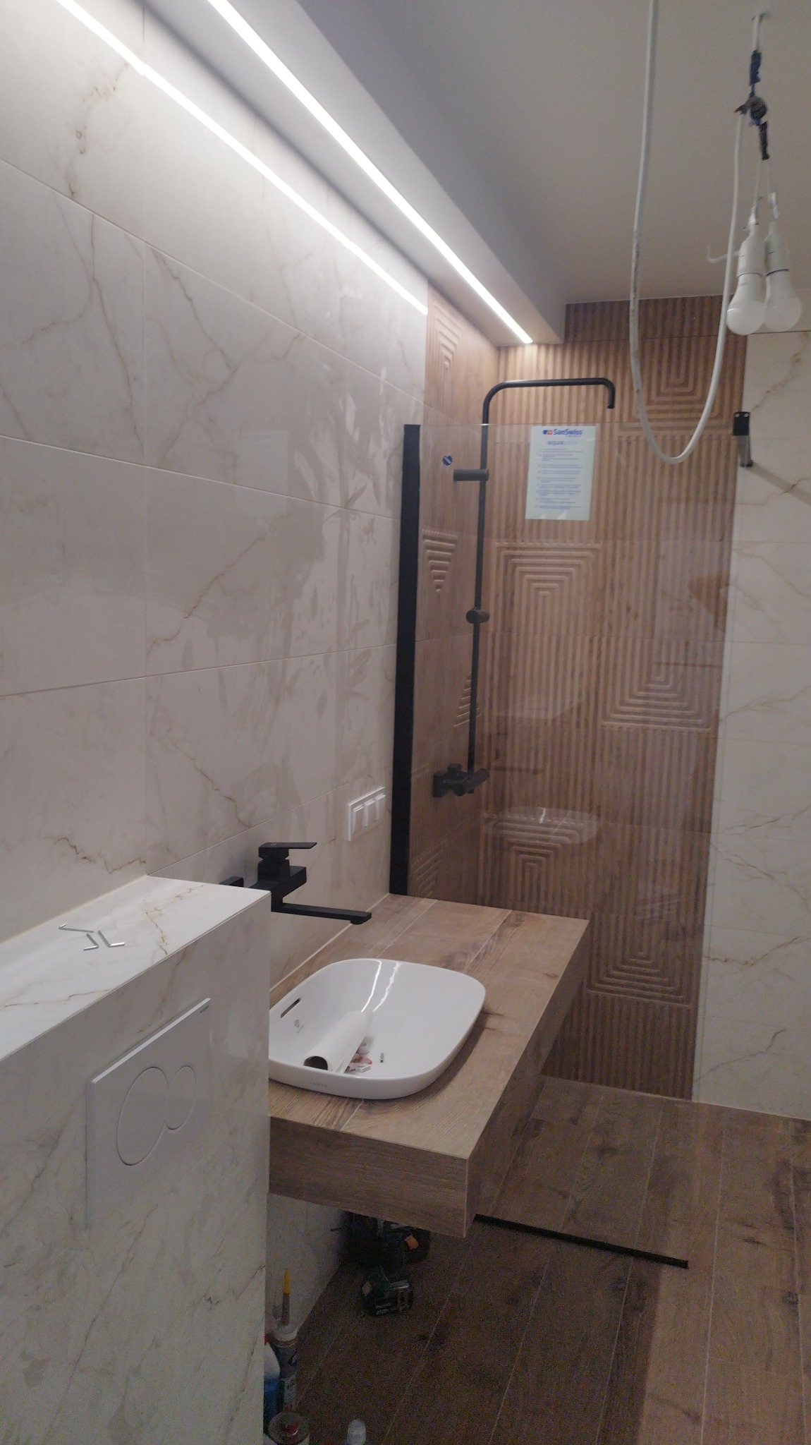 Nowoczesna łazienka z beżowymi płytkami imitującymi marmur, podświetleniem LED, umywalką nablatową na drewnopodobnym blacie, czarną armaturą i kabiną prysznicową z dekoracyjną ścianą. Widoczne...