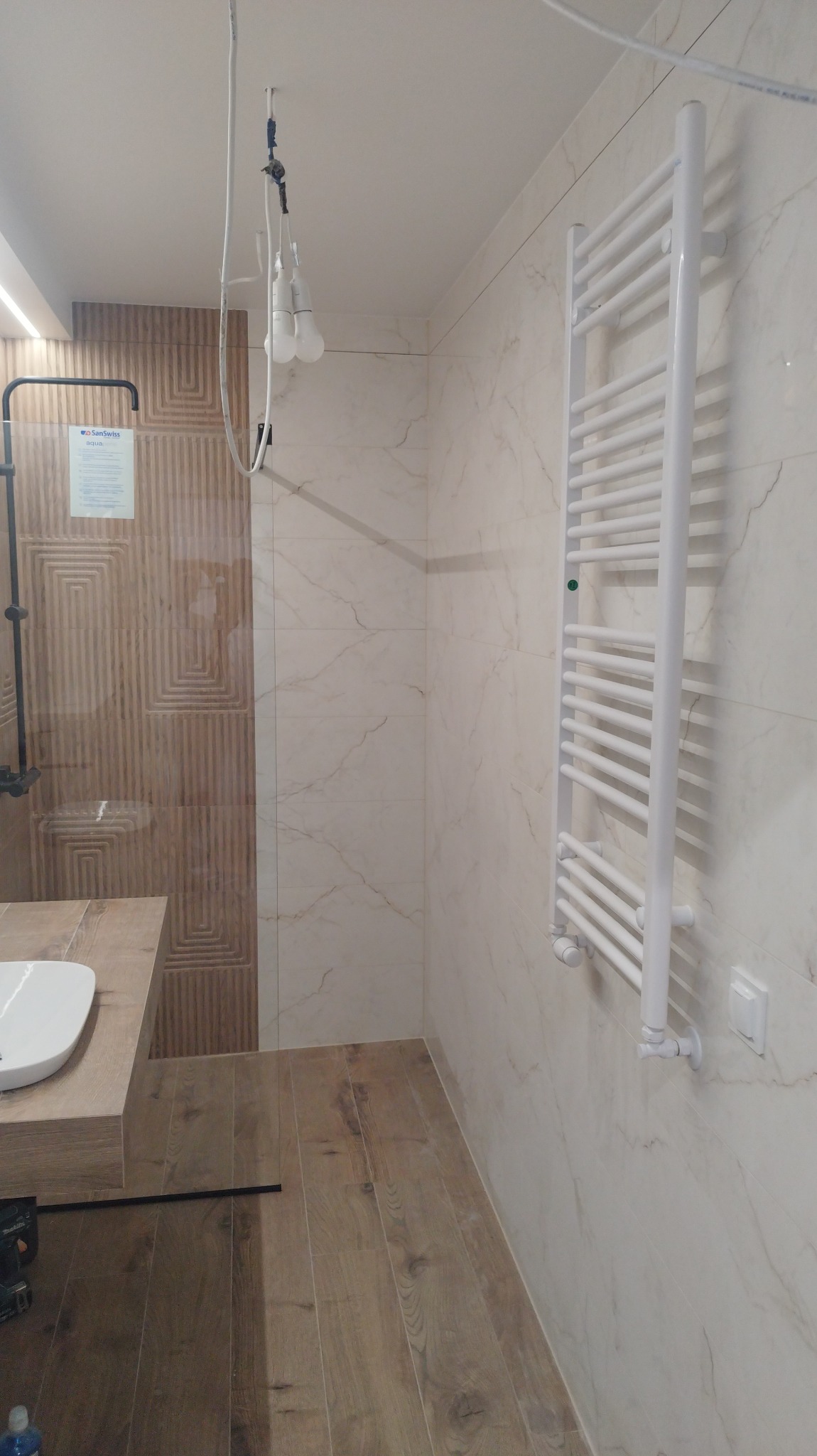 Wnętrze łazienki w trakcie prac wykończeniowych: zamontowany grzejnik drabinkowy, częściowo ułożone płytki imitujące marmur, drewnopodobna podłoga, tymczasowe oświetlenie wiszące na kablach.