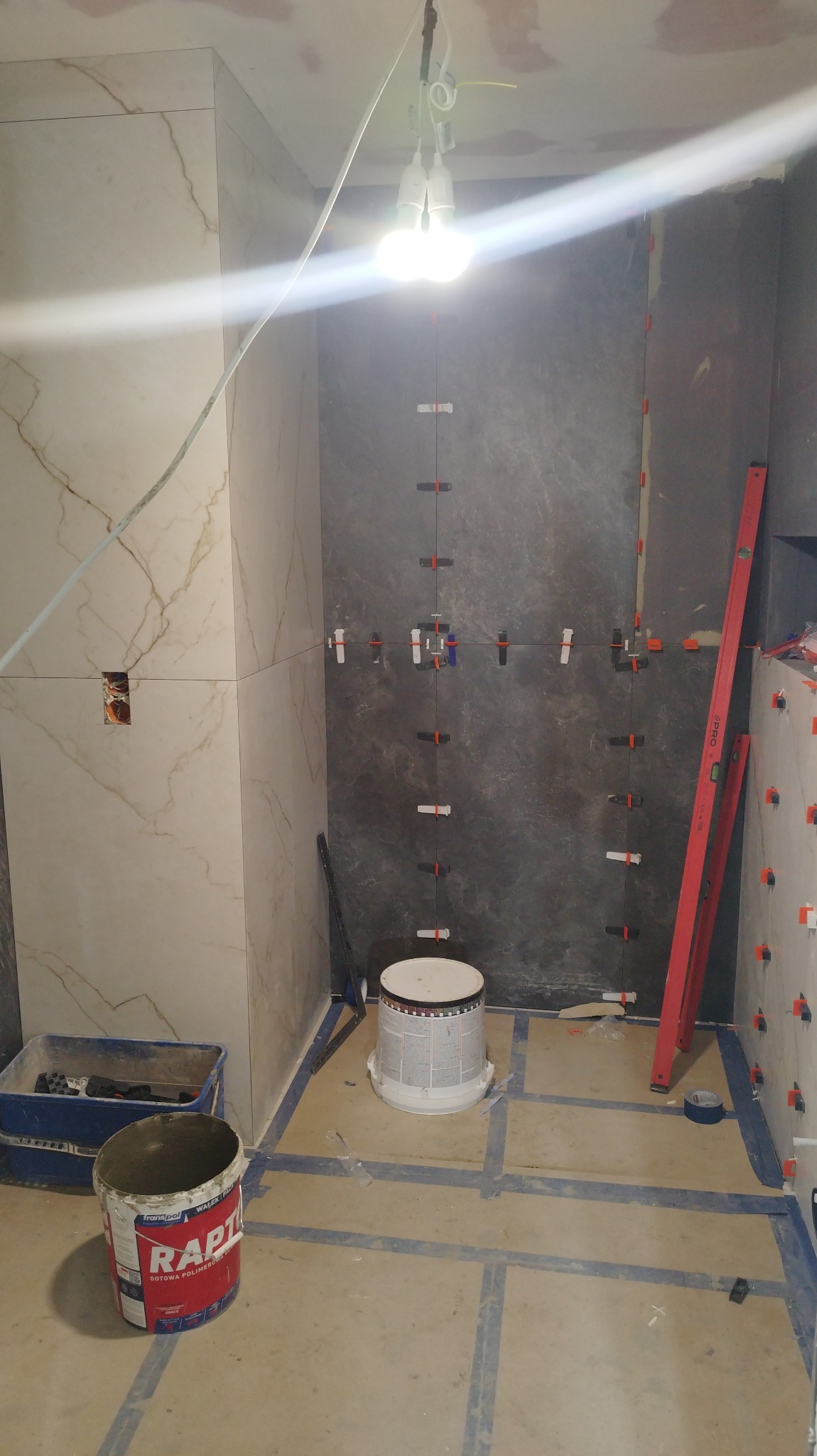 W trakcie układania płytek w łazience: widoczne płytki imitujące marmur na ścianie, ciemne płytki na innej ścianie z krzyżykami poziomującymi, poziomnica oparta o ścianę, wiadro z zaprawą Raptor...