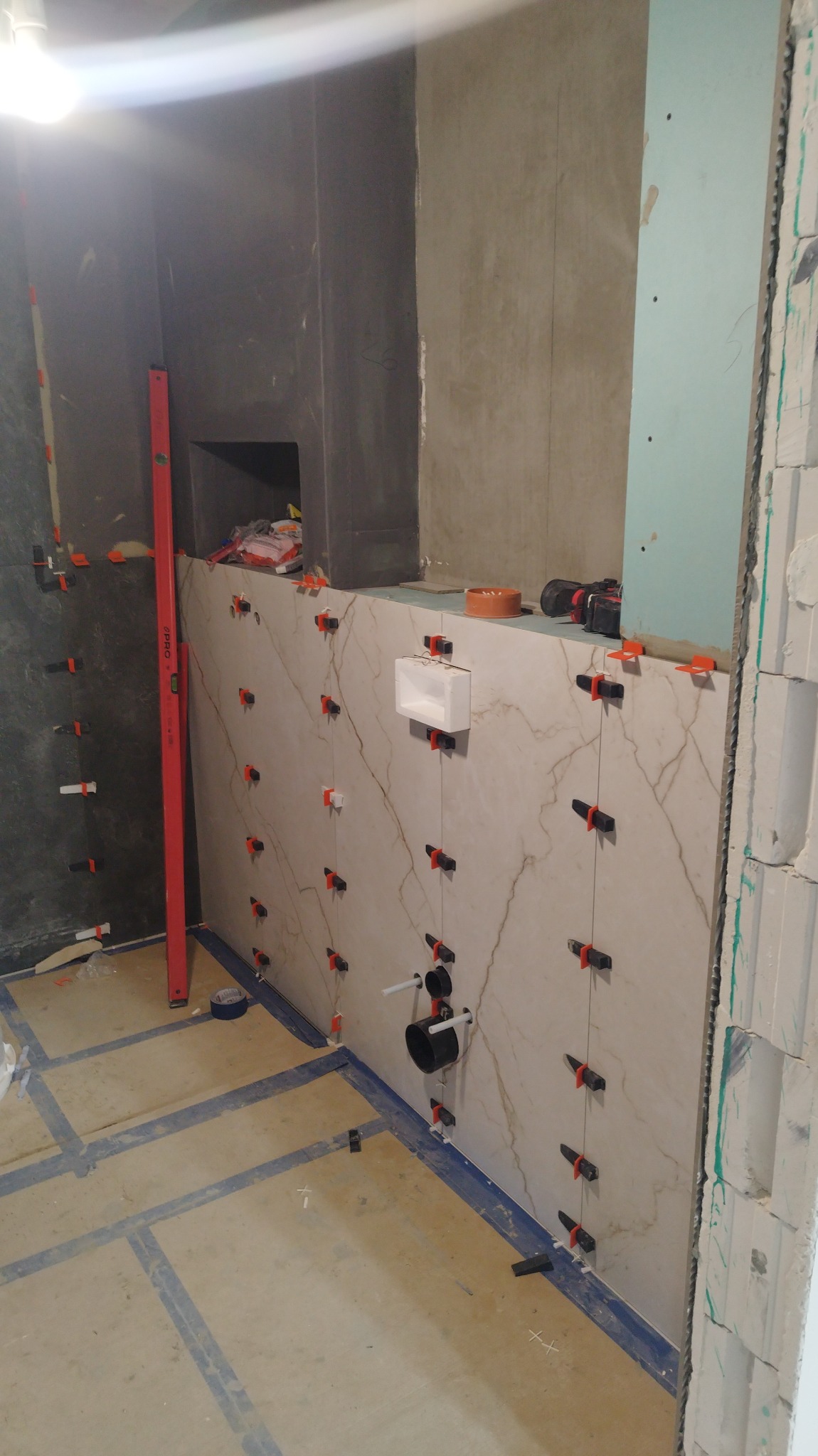 Wnętrze łazienki w trakcie remontu: częściowo wyłożone marmuropodobnymi płytkami z systemem poziomowania, widoczne instalacje wodno-kanalizacyjne, poziomica pionowa przy ścianie, przygotowane...