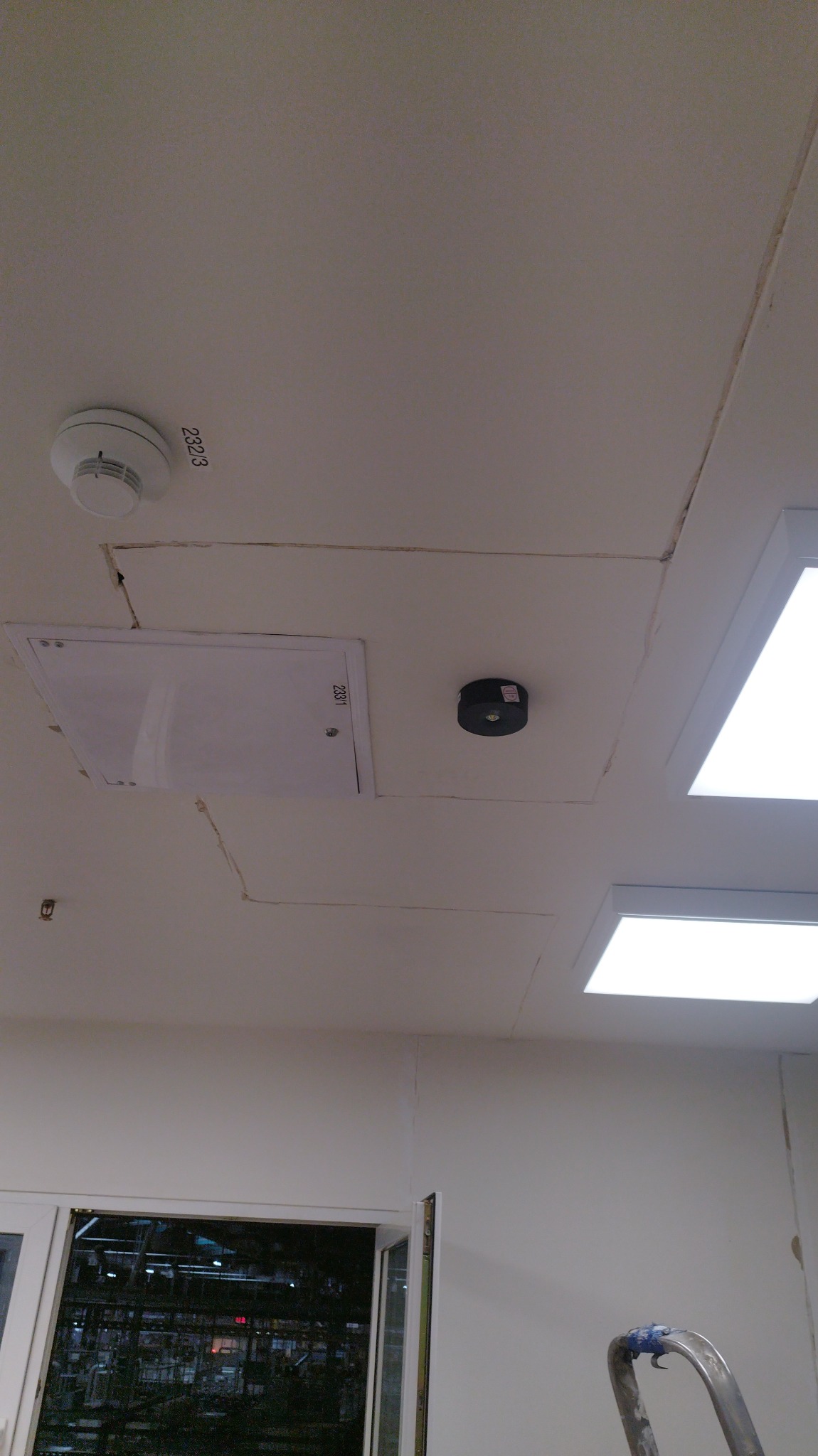 Sufit z widocznymi pęknięciami na łączeniach płyt, czujnik dymu, czujnik ruchu, klapa rewizyjna i lampy LED, widoczne ślady po pracach wykończeniowych