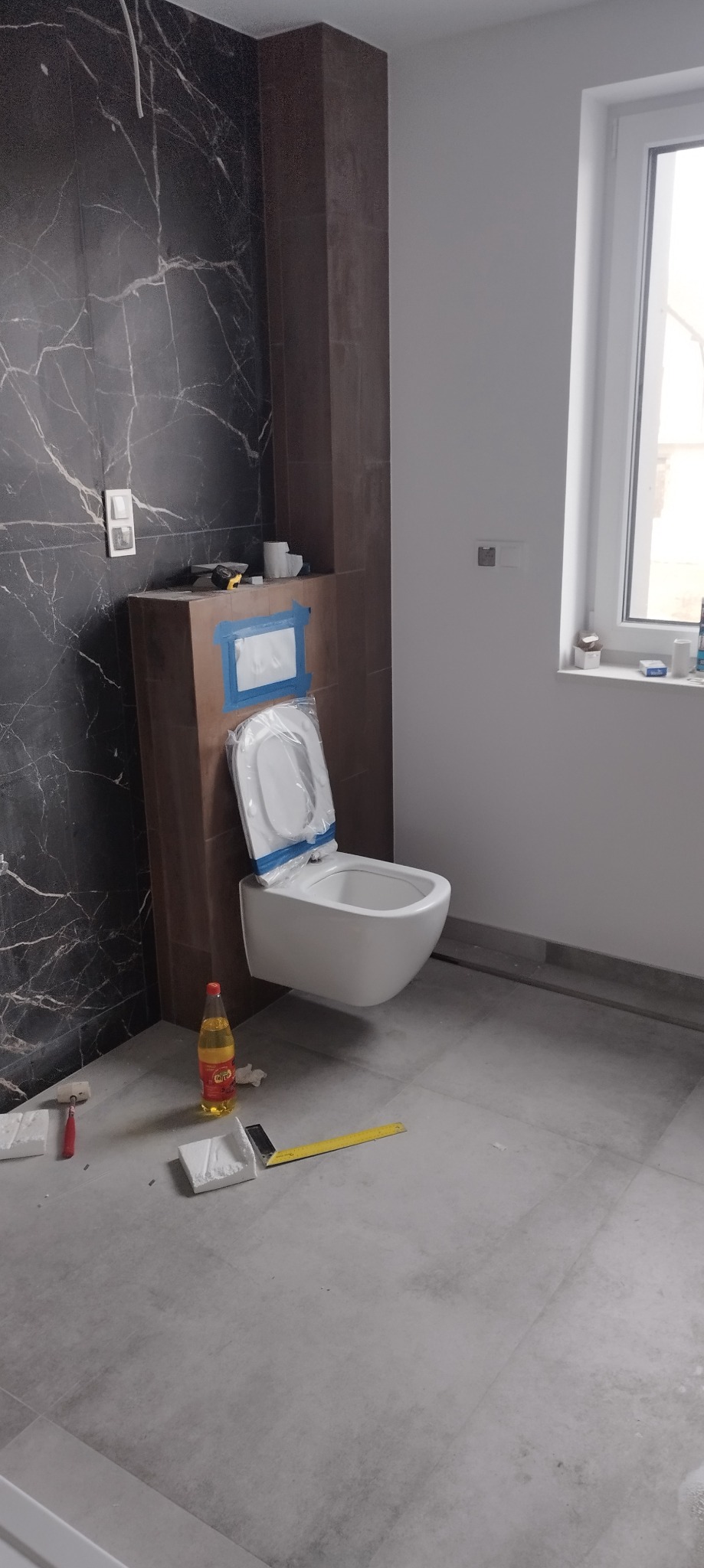 Podwieszana miska WC w trakcie montażu w łazience z marmurową ścianą, widoczne narzędzia i butelka na podłodze.