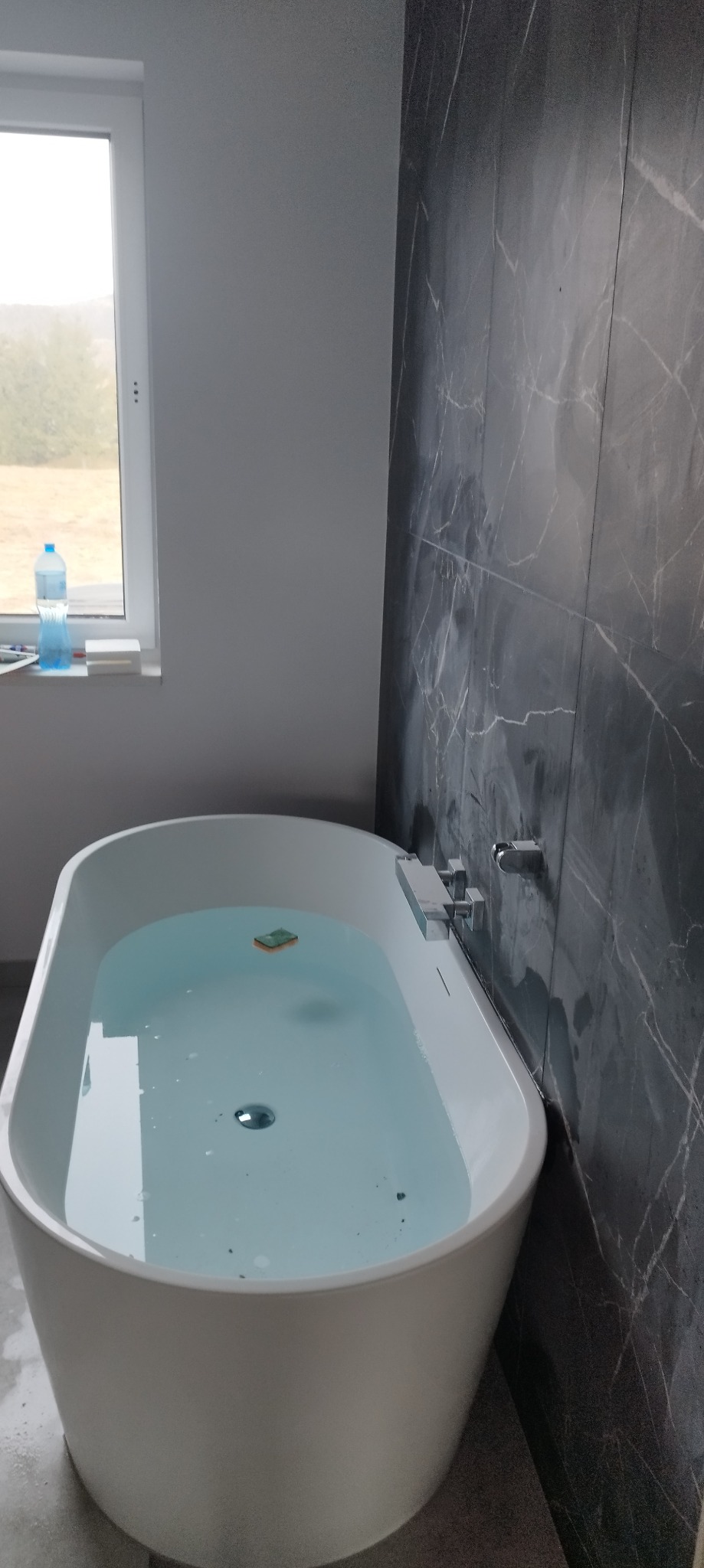 Wanna wolnostojąca wypełniona wodą, z widoczną gąbką na powierzchni, na tle ściany z płytek imitujących marmur i okna z butelką wody na parapecie.