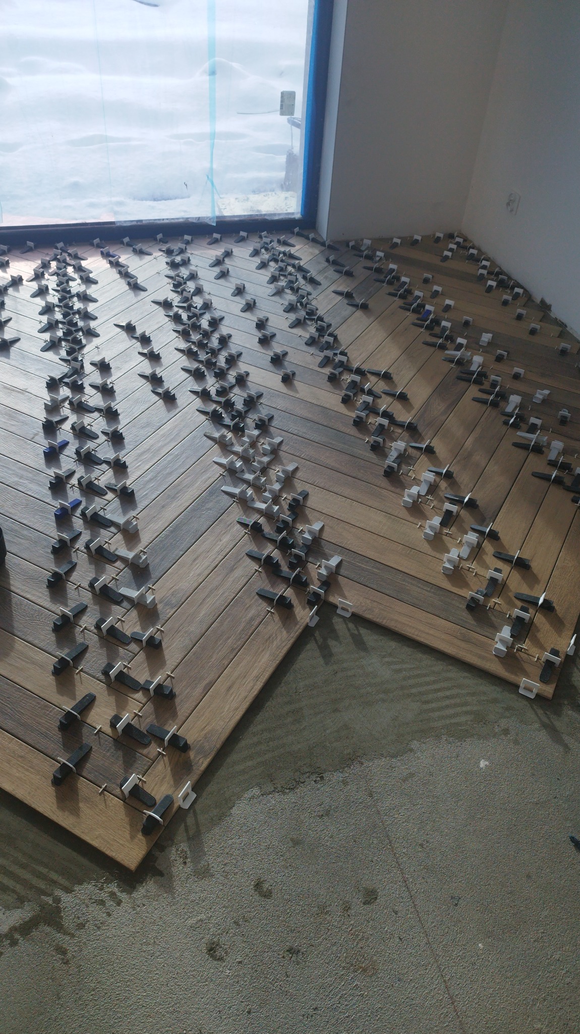 Układanie drewnopodobnych płytek podłogowych w jodełkę, z użyciem klipsów poziomujących, widok na okno z zimowym krajobrazem.