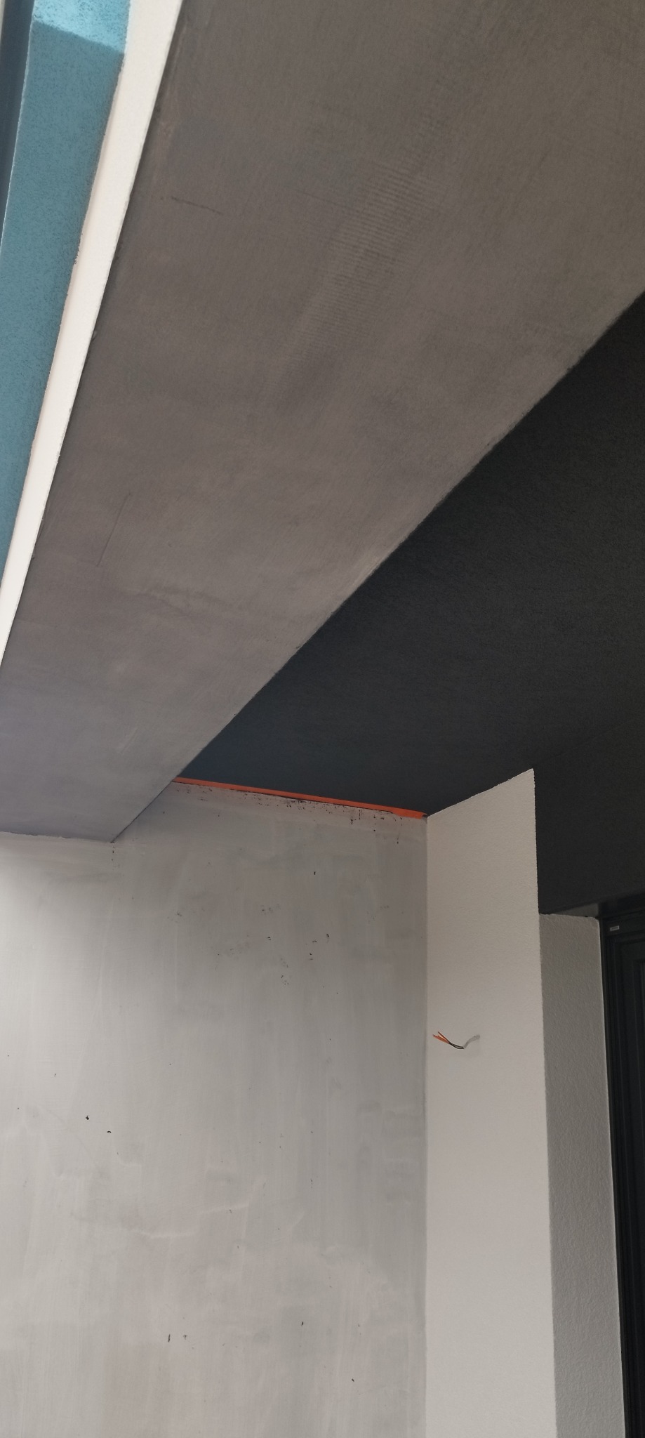 Narożnik pomieszczenia w trakcie prac wykończeniowych: szara gładź sufitowa, surowa ściana przygotowana pod malowanie, wystające przewody elektryczne.