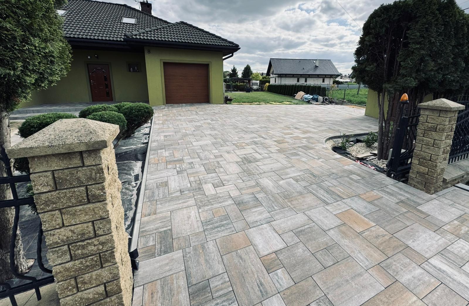 Ułożona kostka brukowa w różnych odcieniach szarości i beżu tworzy wzór na podjeździe przed domem. Widoczne ogrodzenie z kamiennymi słupkami i zieleń ogrodu.