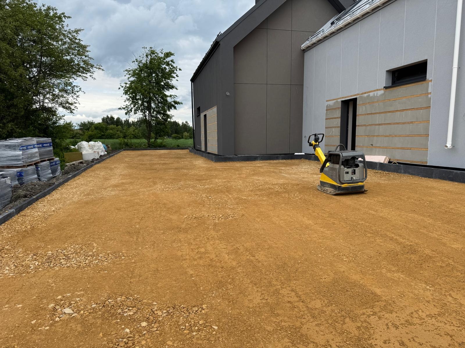 Przygotowany teren pod kostkę brukową z użyciem zagęszczarki. Widoczne obrzeża, podsypka i palety kostki brukowej w tle.