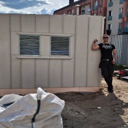 Dach-Felc - Niewielki, beżowy budynek z dwoma oknami z żaluzjami, na tle błękitnego nieba z chmurami. Obok stoi mężczyzna w czarnym ubraniu.