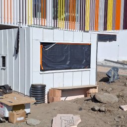 Dach-Felc - Biała, modułowa konstrukcja w trakcie budowy, z widocznym oknem zabezpieczonym folią. W tle budynek z kolorowymi panelami i pracownik z telefonem. Plac budowy w słoneczny dzień.