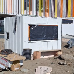 Dach-Felc - Budowa małego, białego budynku z blachy falistej na placu budowy. Widoczny pracownik w oddali i materiały budowlane. Otwór okienny zabezpieczony czarną folią.