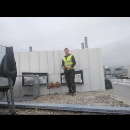 Dach-Felc - Pracownik w kamizelce na dachu z blachy trapezowej, obok kominy wentylacyjne. Widoczne elementy instalacji i konstrukcji dachowej.