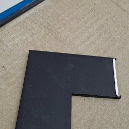 Dach-Felc - Czarny, kątowy element blaszany z metalowymi krawędziami, leżący na betonowym podłożu, prawdopodobnie fragment obróbki blacharskiej dachu.