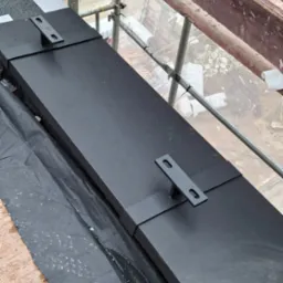 Czarny pas obróbki blacharskiej z uchwytami na dachu, widok z góry z rusztowania. Czarna membrana dachowa i płyta OSB widoczne. Detal obróbki.