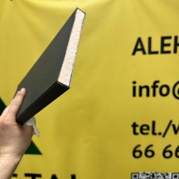 Dach-Felc - Przekrój panelu ogrodzeniowego w dłoni na tle żółtego baneru reklamowego firmy. Widoczna struktura wewnętrzna panelu.