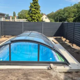 Nowoczesne ogrodzenie panelowe w kolorze grafitowym otacza basen z zadaszeniem. Widoczne elementy konstrukcyjne ogrodzenia i betonowa podstawa basenu.