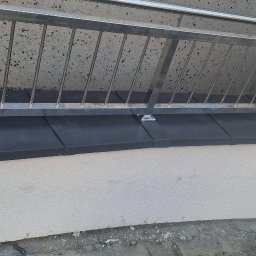 Dach-Felc - Nowoczesna, metalowa balustrada okienna z czarnym parapetem, zamontowana na elewacji budynku o jasnej, chropowatej strukturze. Minimalistyczny design i precyzyjne wykonanie.