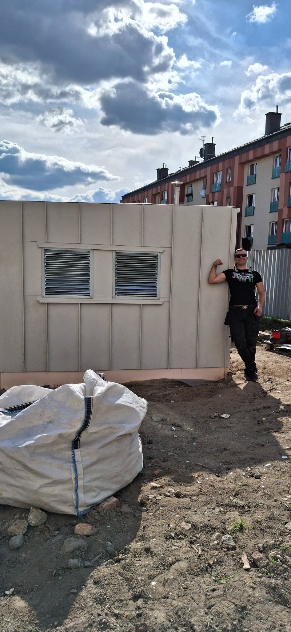 Niewielki, beżowy budynek z dwoma oknami z żaluzjami, na tle błękitnego nieba z chmurami. Obok stoi mężczyzna w czarnym ubraniu.