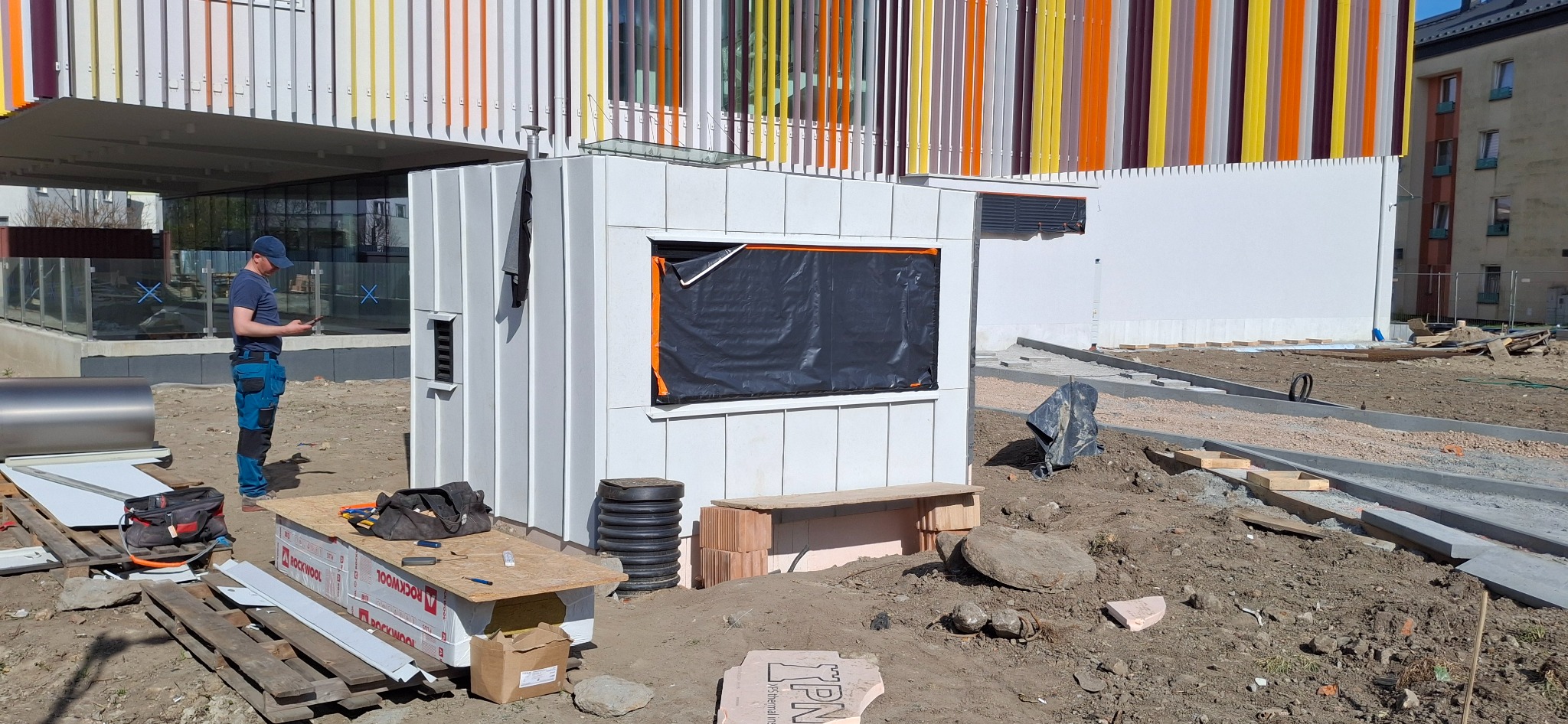 Biała, modułowa konstrukcja w trakcie budowy, z widocznym oknem zabezpieczonym folią. W tle budynek z kolorowymi panelami i pracownik z telefonem. Plac budowy w słoneczny dzień.