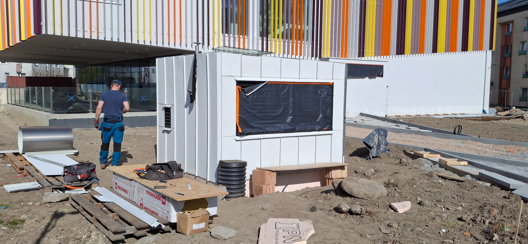 Budowa małego, białego budynku z blachy falistej na placu budowy. Widoczny pracownik w oddali i materiały budowlane. Otwór okienny zabezpieczony czarną folią.