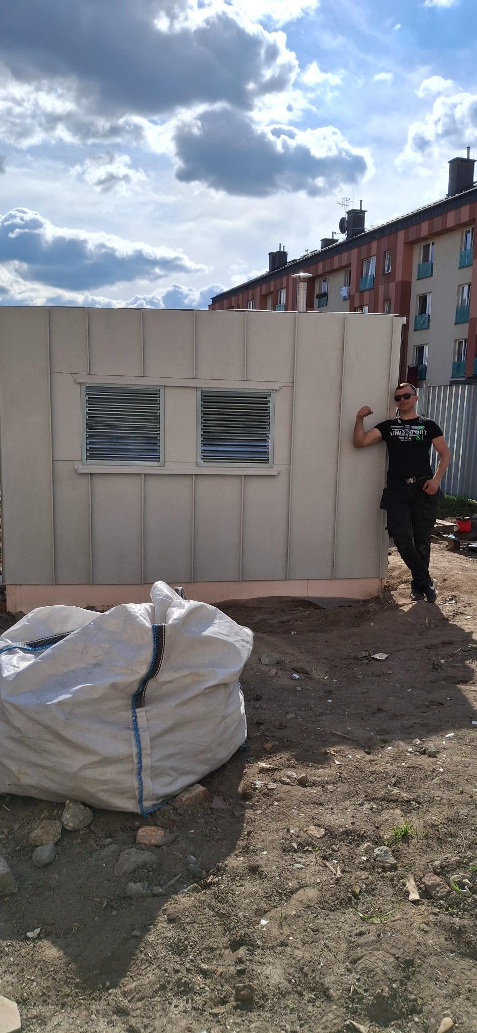 Szara budka z blachy trapezowej z oknami wentylacyjnymi, pracownik oparty o ścianę, worek z odpadami budowlanymi na ziemi, w tle budynek mieszkalny i pochmurne niebo.