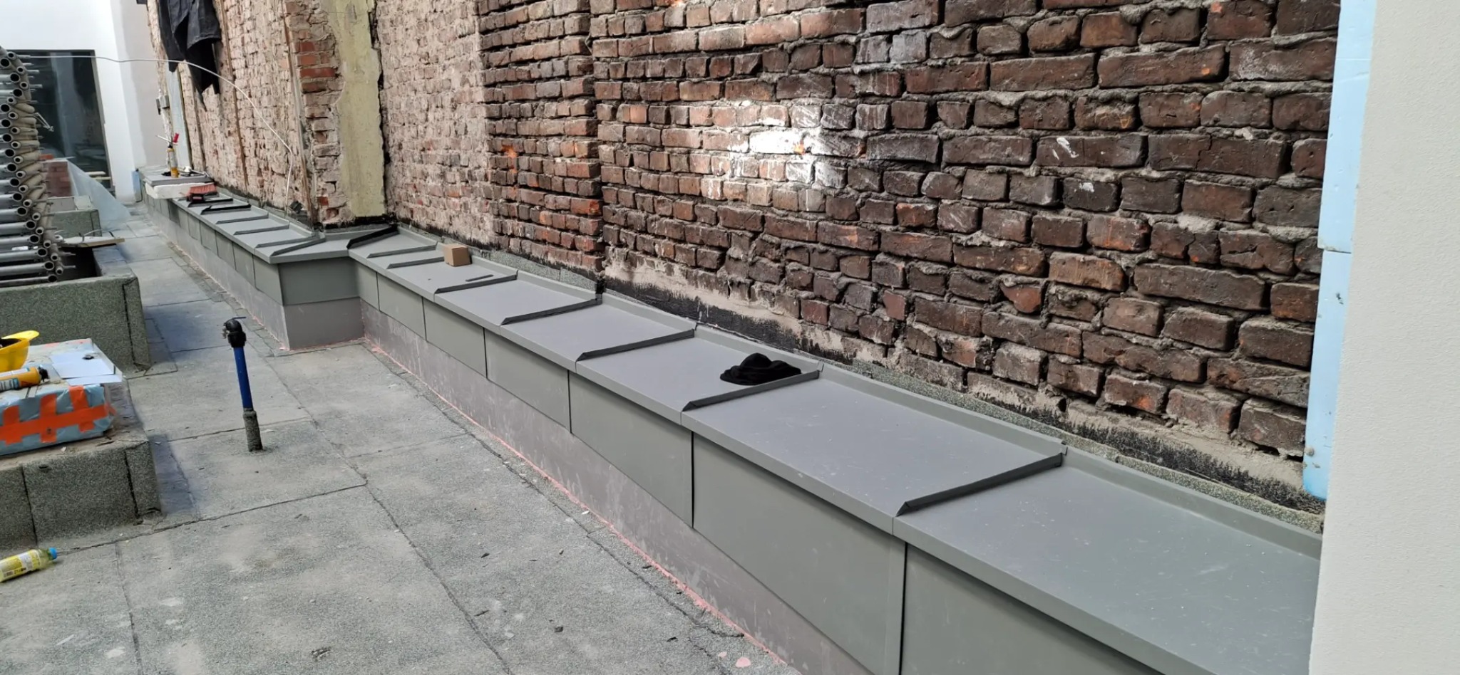 Szara, metalowa obróbka blacharska przy ceglanej ścianie w Krakowie. Widoczne detale łączeń i kształtów. Praca dekarska w trakcie realizacji.