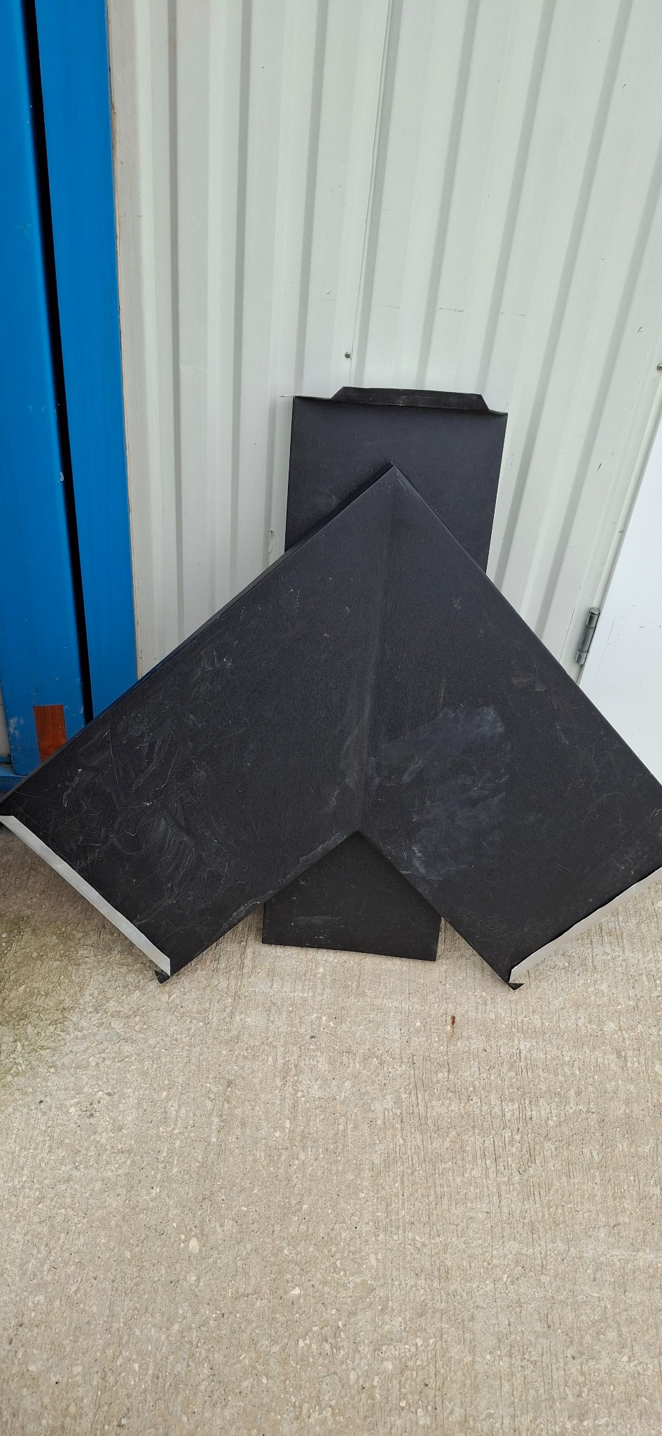 Czarne elementy blaszane, prawdopodobnie obróbki dachowe, leżące na betonowej powierzchni. Widoczne ślady użytkowania i zarysowania.