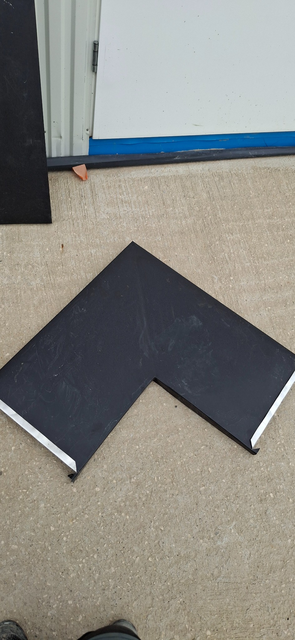 Czarny, kątowy element blaszany z widoczną krawędzią wykończeniową leży na szarej powierzchni. Fragment blachy i drzwi w tle.