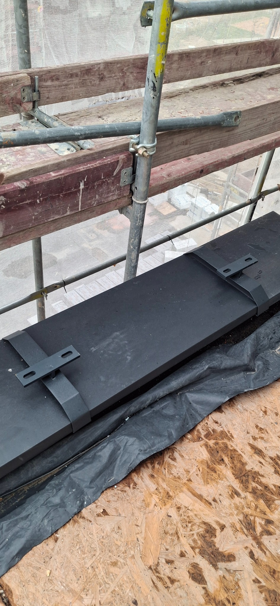 Czarne, metalowe elementy obróbki blacharskiej z uchwytami na drewnianym rusztowaniu, gotowe do montażu na dachu, widok z góry na plac budowy.