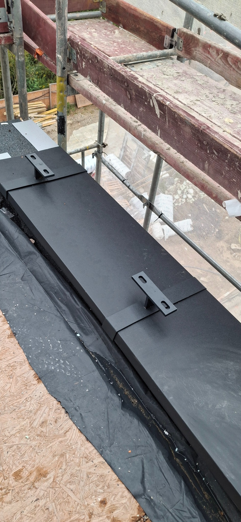 Czarny pas obróbki blacharskiej z uchwytami na dachu, widok z góry z rusztowania. Czarna membrana dachowa i płyta OSB widoczne. Detal obróbki.