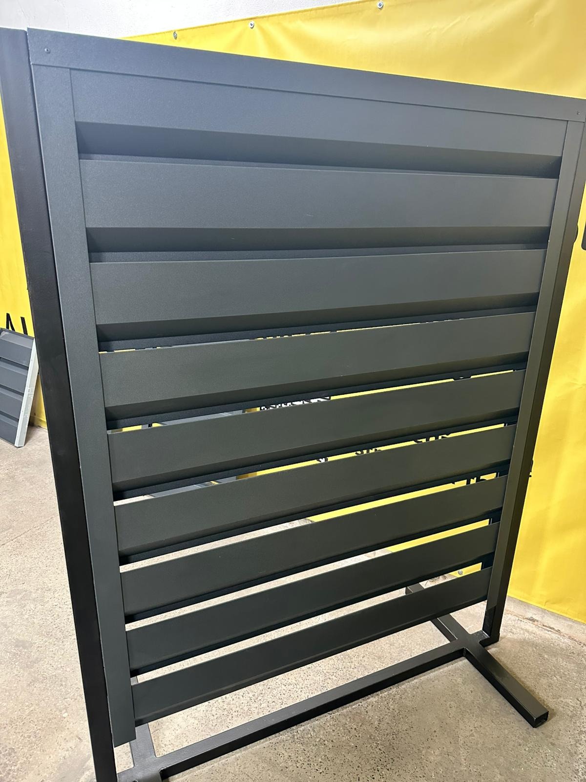 Grafitowy panel ogrodzeniowy z poziomych listew, widoczny z bliska na tle żółtej tkaniny. Konstrukcja metalowa z wyraźnymi łączeniami.