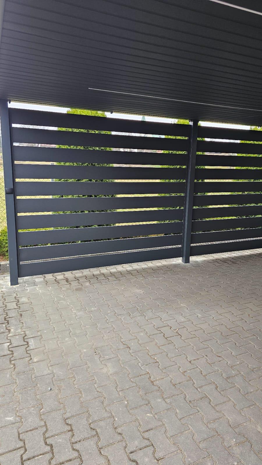 Nowoczesne, antracytowe ogrodzenie panelowe z poziomymi szczeblami, zamontowane pod zadaszeniem, widok z zewnątrz na kostkę brukową.