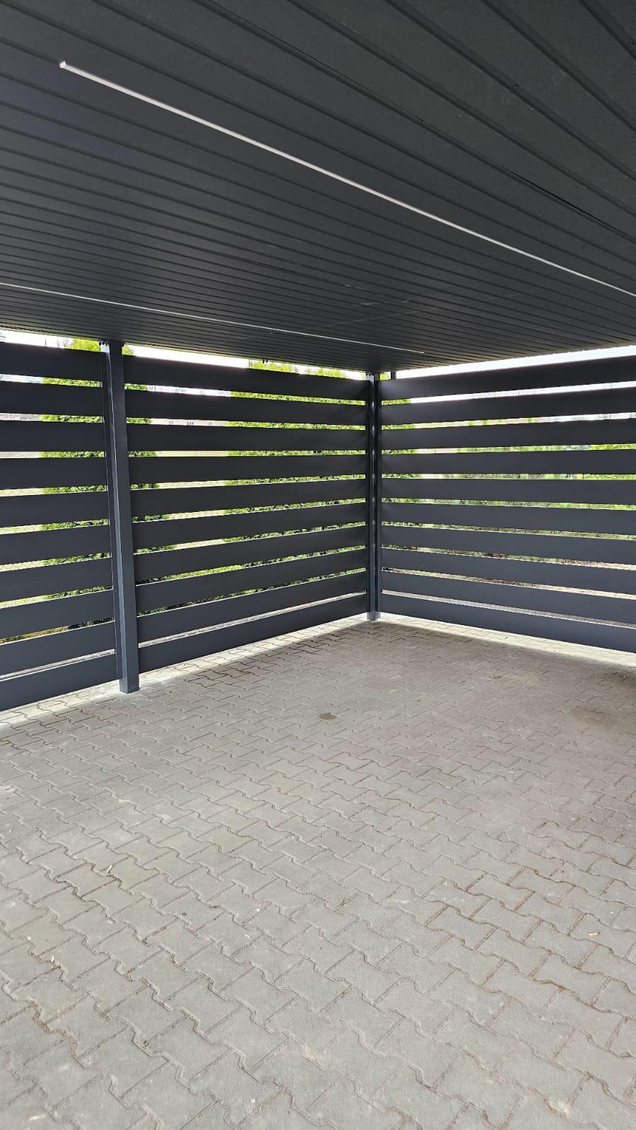 Nowoczesna, minimalistyczna wiata garażowa z poziomych, grafitowych paneli metalowych, wykończona kostką brukową. Widok z wnętrza, na zewnątrz zieleń.