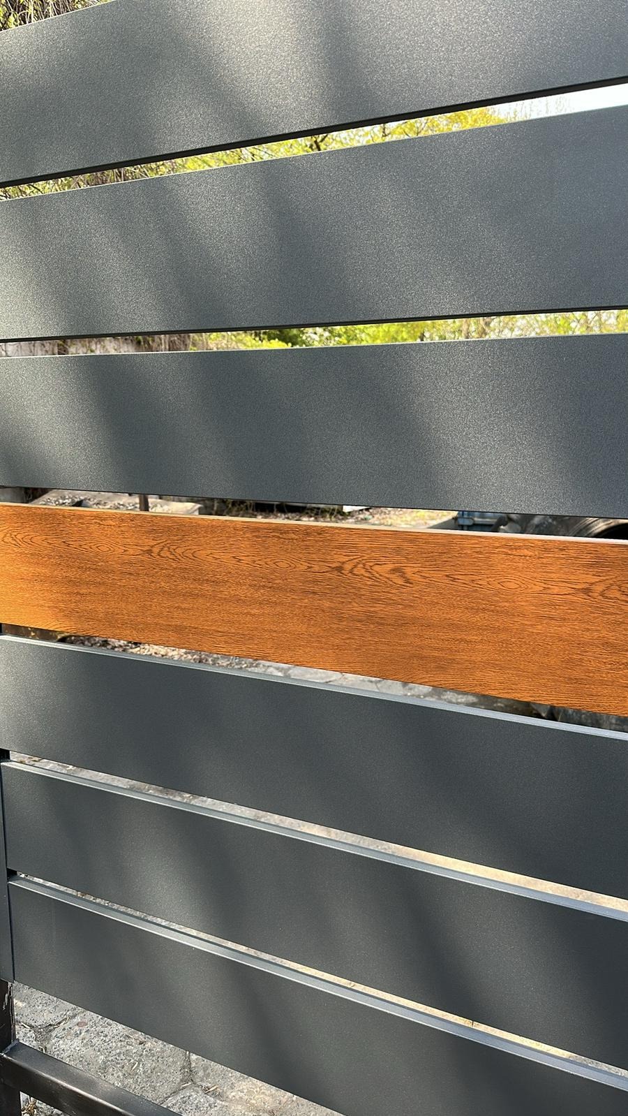 Nowoczesne ogrodzenie panelowe z poziomych, szarych profili z jednym akcentem w kolorze drewna, widoczne z bliska, z zielenią w tle.