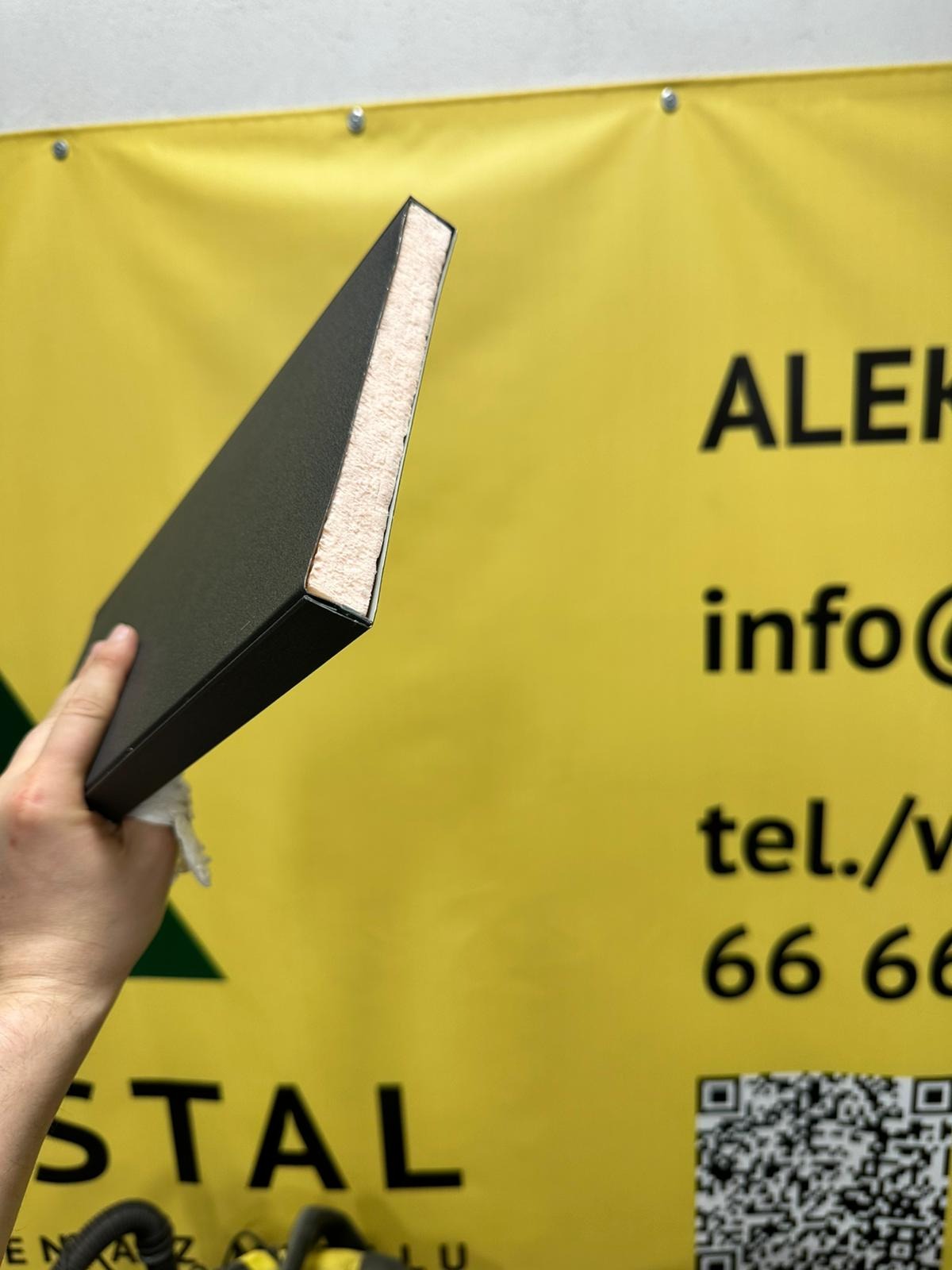 Przekrój panelu ogrodzeniowego w dłoni na tle żółtego baneru reklamowego firmy. Widoczna struktura wewnętrzna panelu.