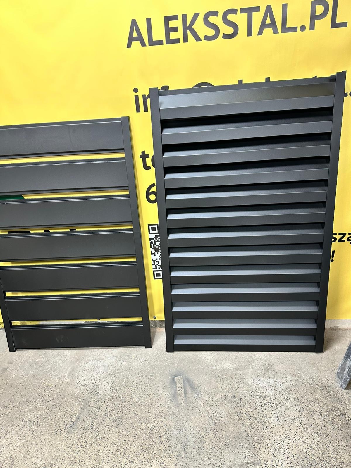 Dwa panele ogrodzeniowe metalowe, czarne, poziome lamele, na szarym tle posadzki i żółtym tle z logo. Widoczny kod QR na jednym z paneli.