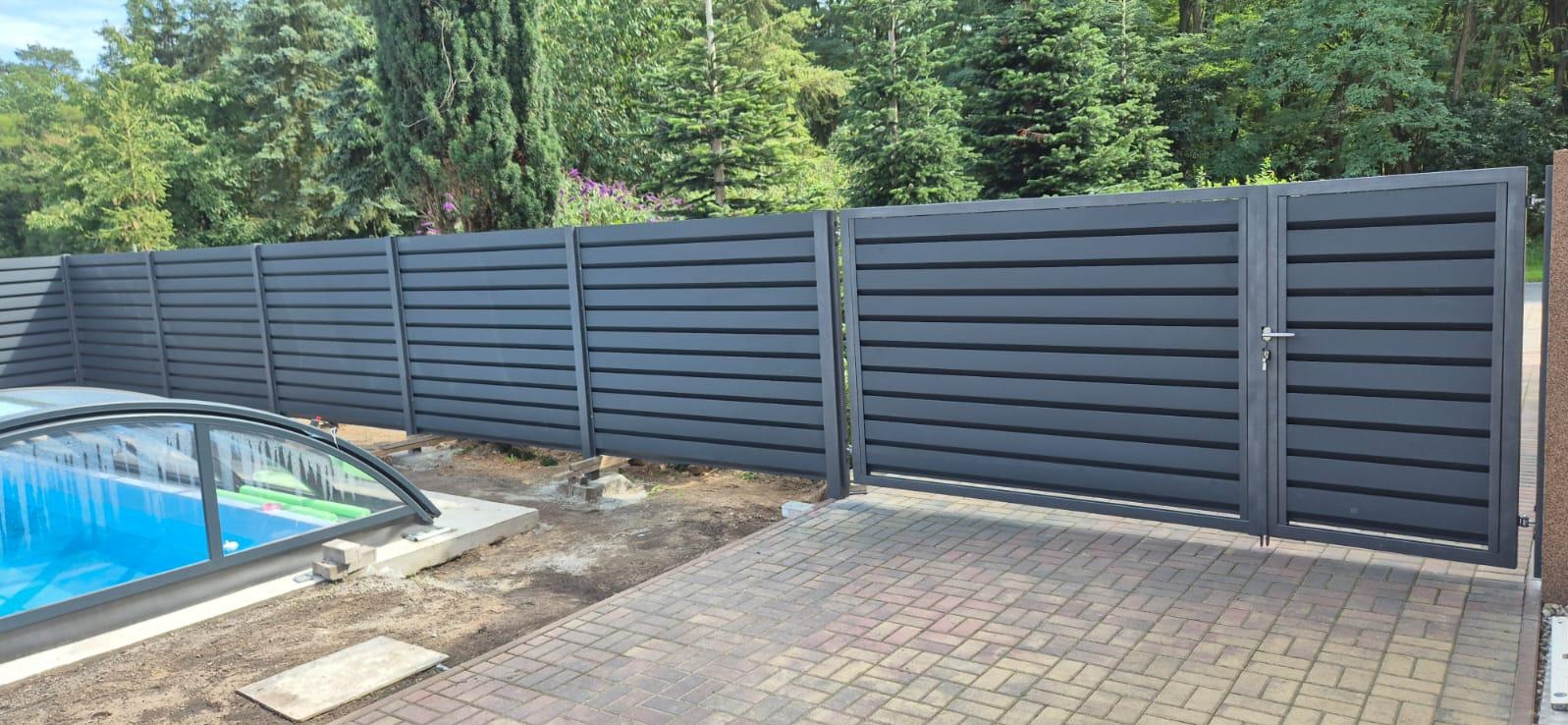 Nowoczesne, poziome ogrodzenie panelowe w kolorze grafitowym z bramą, przylegające do posesji z basenem i brukowanym podjazdem, otoczone zielenią drzew.