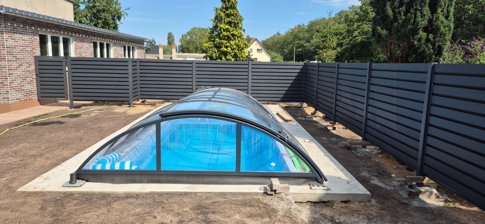 Nowoczesne ogrodzenie panelowe w kolorze grafitowym otacza basen z zadaszeniem. Widoczne elementy konstrukcyjne ogrodzenia i betonowa podstawa basenu.