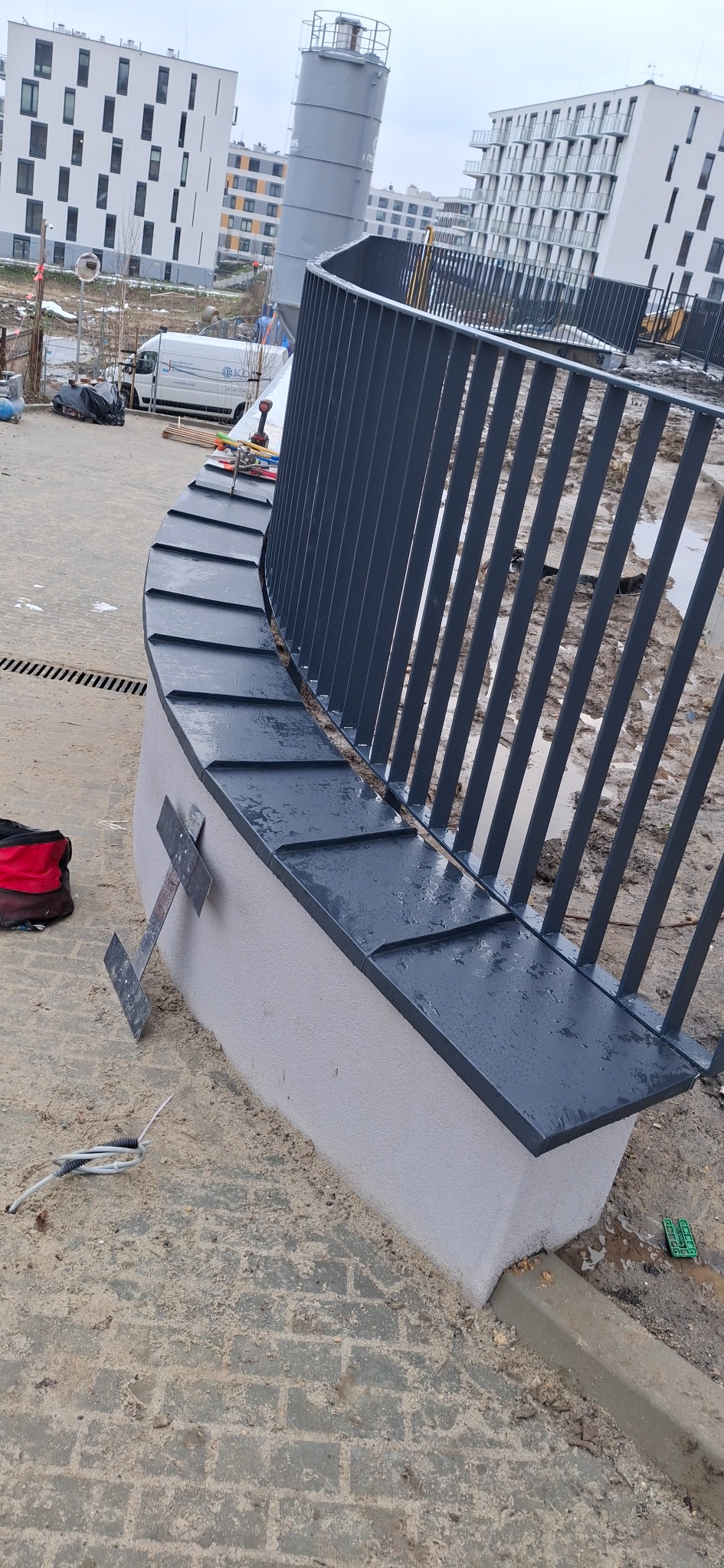 Zakrzywiona balustrada z metalowymi prętami i obróbką blacharską na betonowej podstawie. W tle budynki mieszkalne i elementy placu budowy.