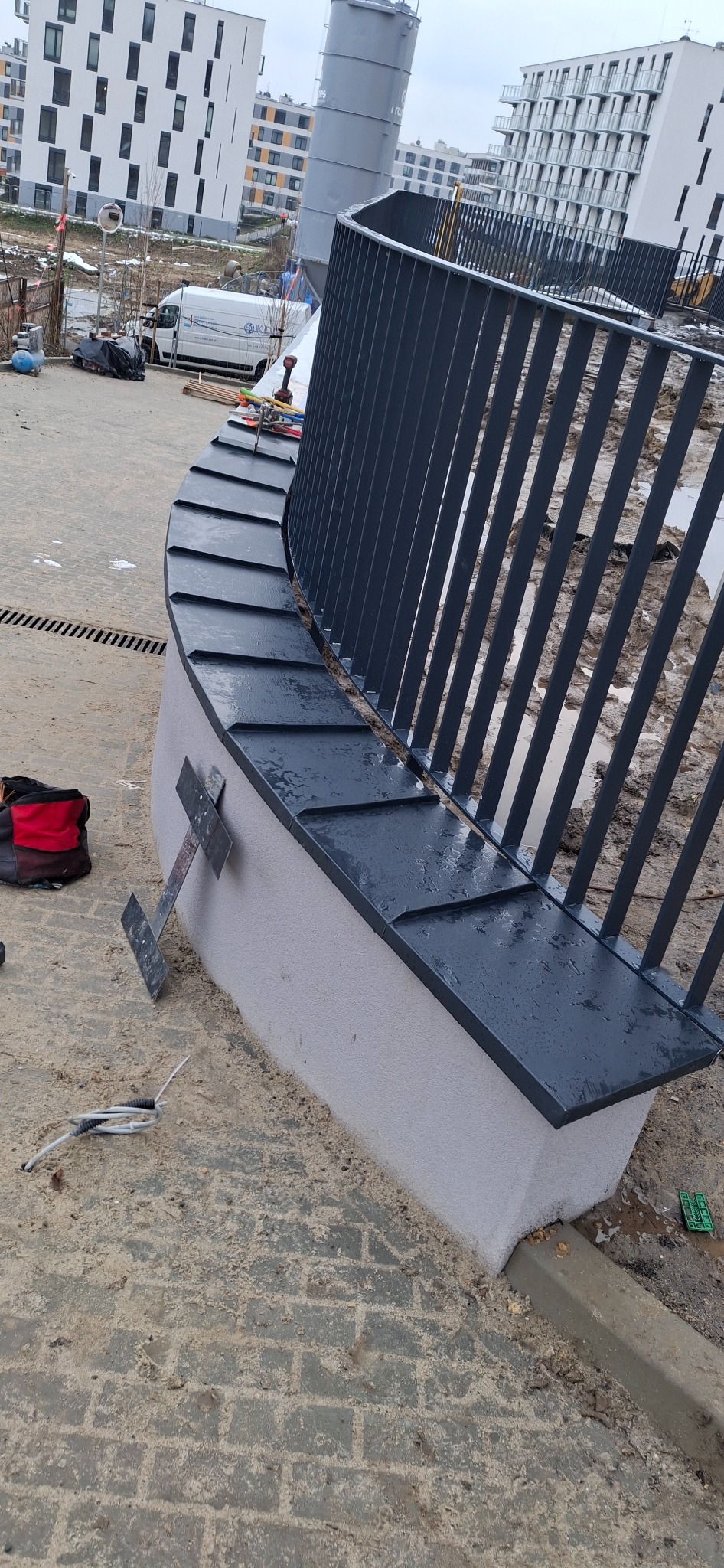 Zakrzywione, szare ogrodzenie metalowe z blacharskim wykończeniem na murku, na tle budynków mieszkalnych. Widoczne narzędzia i elementy montażowe.