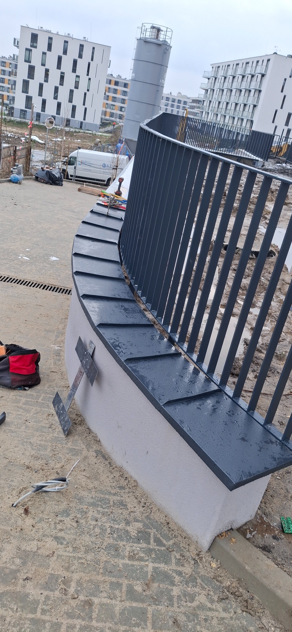 Szara, metalowa balustrada z obróbką blacharską na murku, na tle zabudowy miejskiej. Widoczne narzędzia i elementy montażowe. Ujęcie z bliska, podkreślające detale.