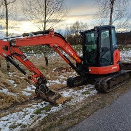 Nadzór i Budownictwo Adam Dyba - Pomarańczowa minikoparka Kubota U48-4 na skraju asfaltowej drogi i zaśnieżonego terenu, z widocznymi śladami gąsienic i łyżką skierowaną w dół.