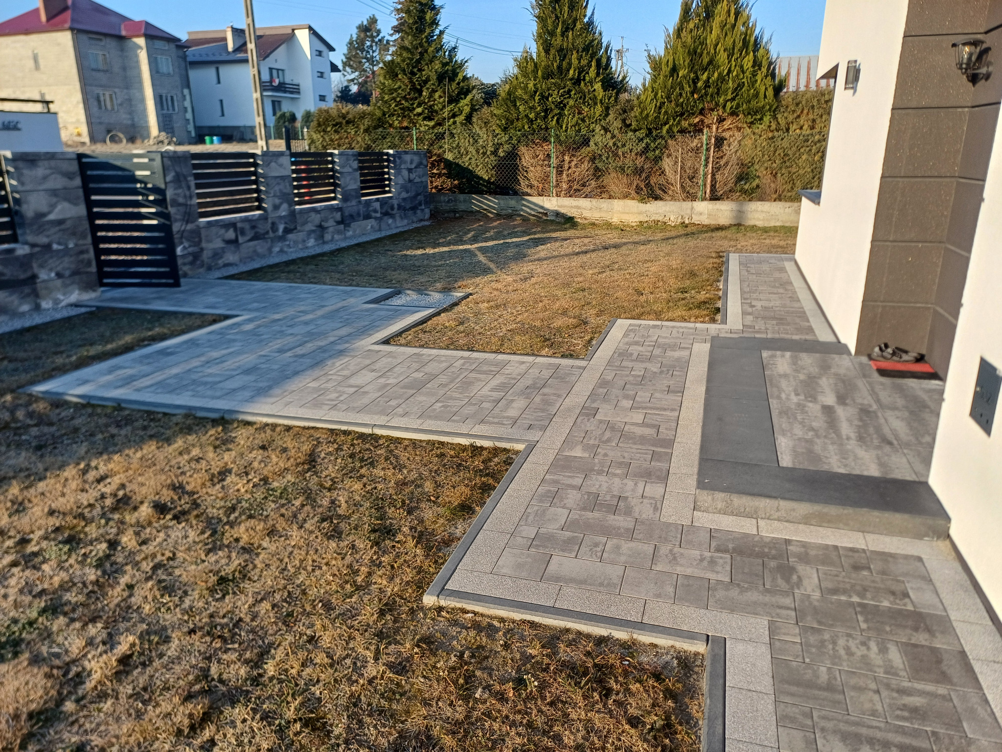 Nowoczesna posesja z szarą kostką brukową tworzącą ścieżkę i podjazd, z minimalistycznym ogrodzeniem z metalowymi przęsłami i elementami z szarego kamienia, na tle trawnika i budynku mieszkalnego.