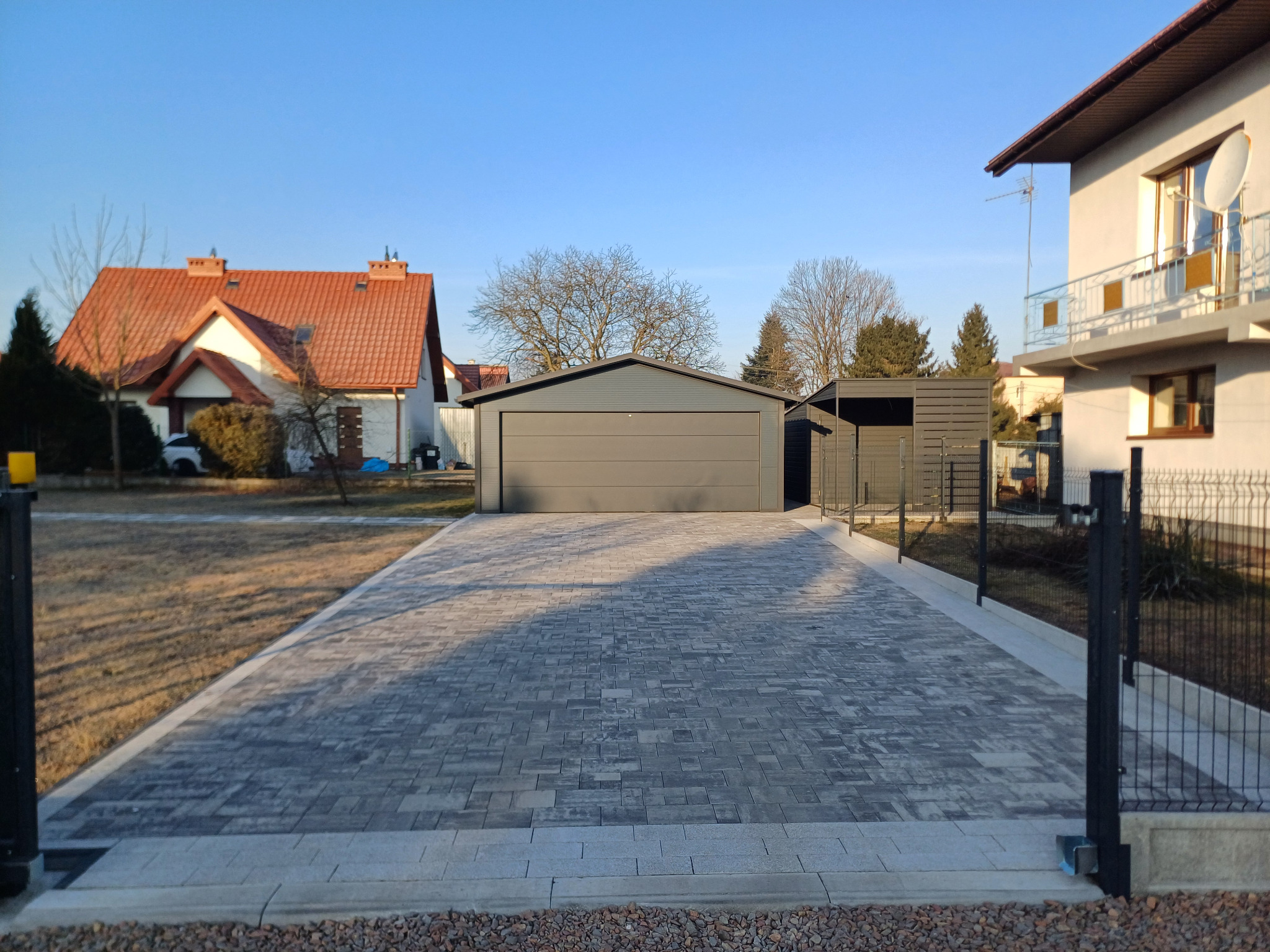 Szeroki podjazd z szarej kostki brukowej prowadzący do garażu blaszanego, widok zza czarnego ogrodzenia w pogodny dzień.