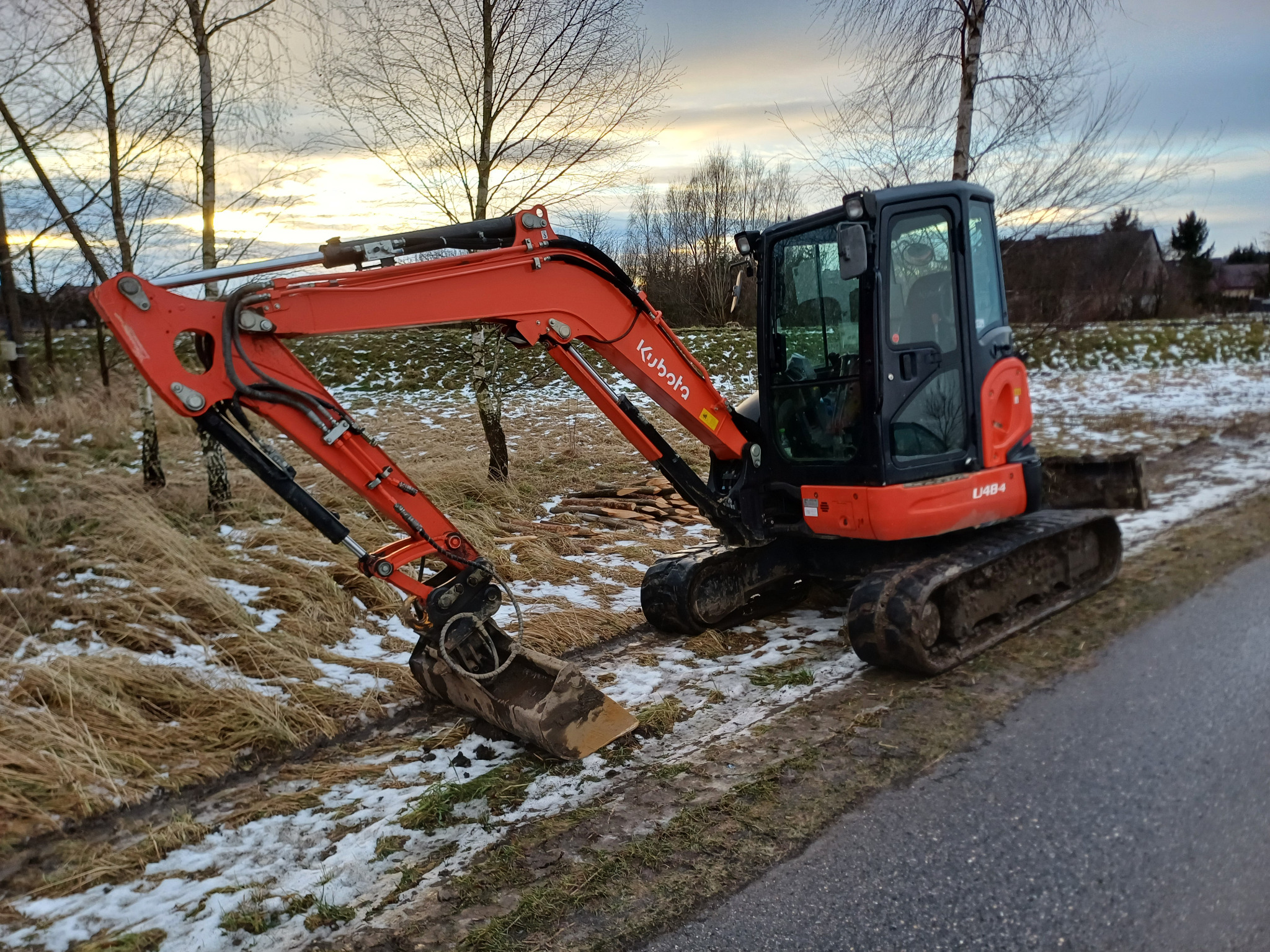 Pomarańczowa minikoparka Kubota U48-4 na skraju asfaltowej drogi i zaśnieżonego terenu, z widocznymi śladami gąsienic i łyżką skierowaną w dół.