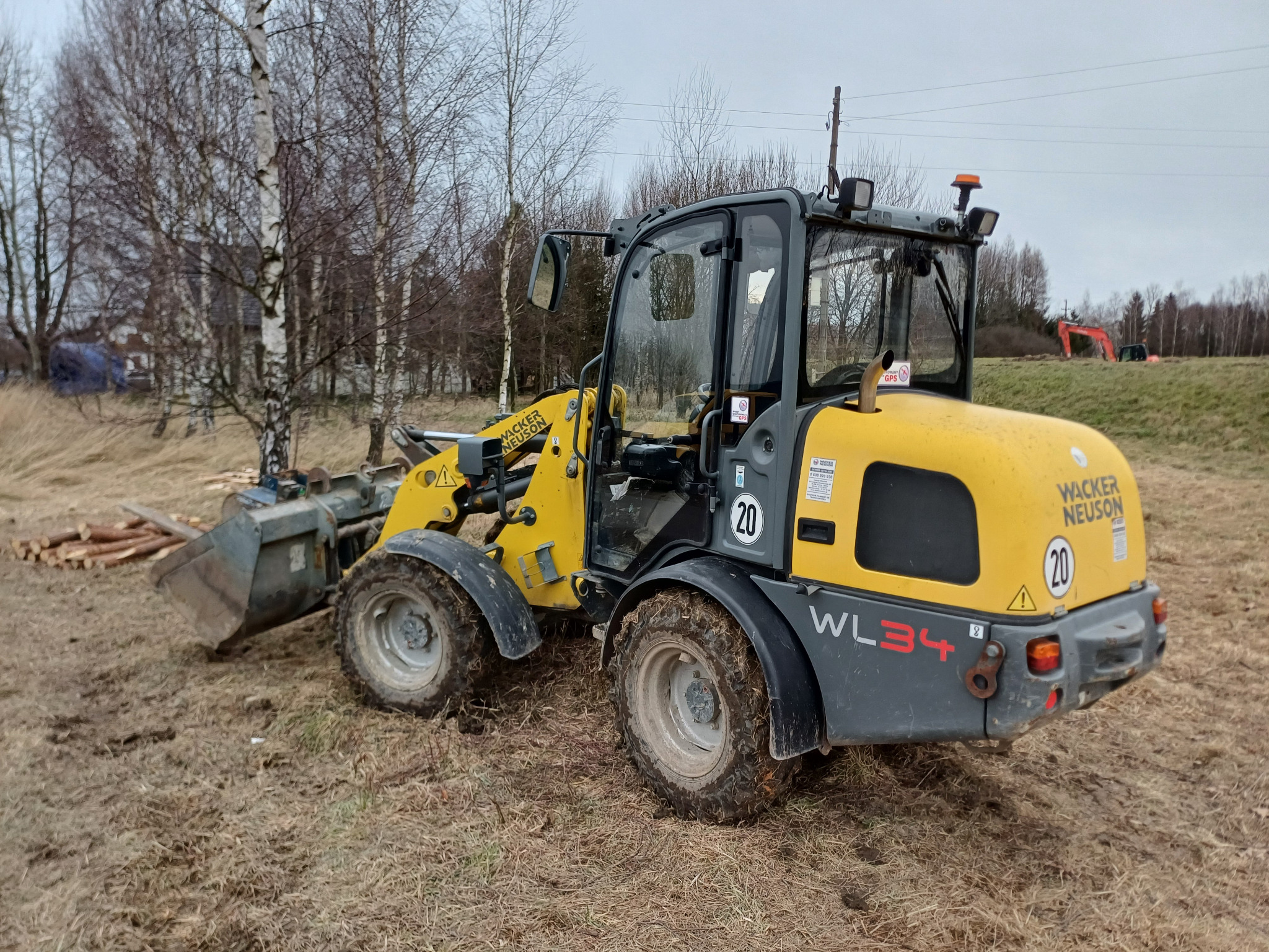 Żółto-szara ładowarka Wacker Neuson WL34 na grząskim terenie z widocznym stosem ściętych gałęzi i w oddali koparka na wzniesieniu.
