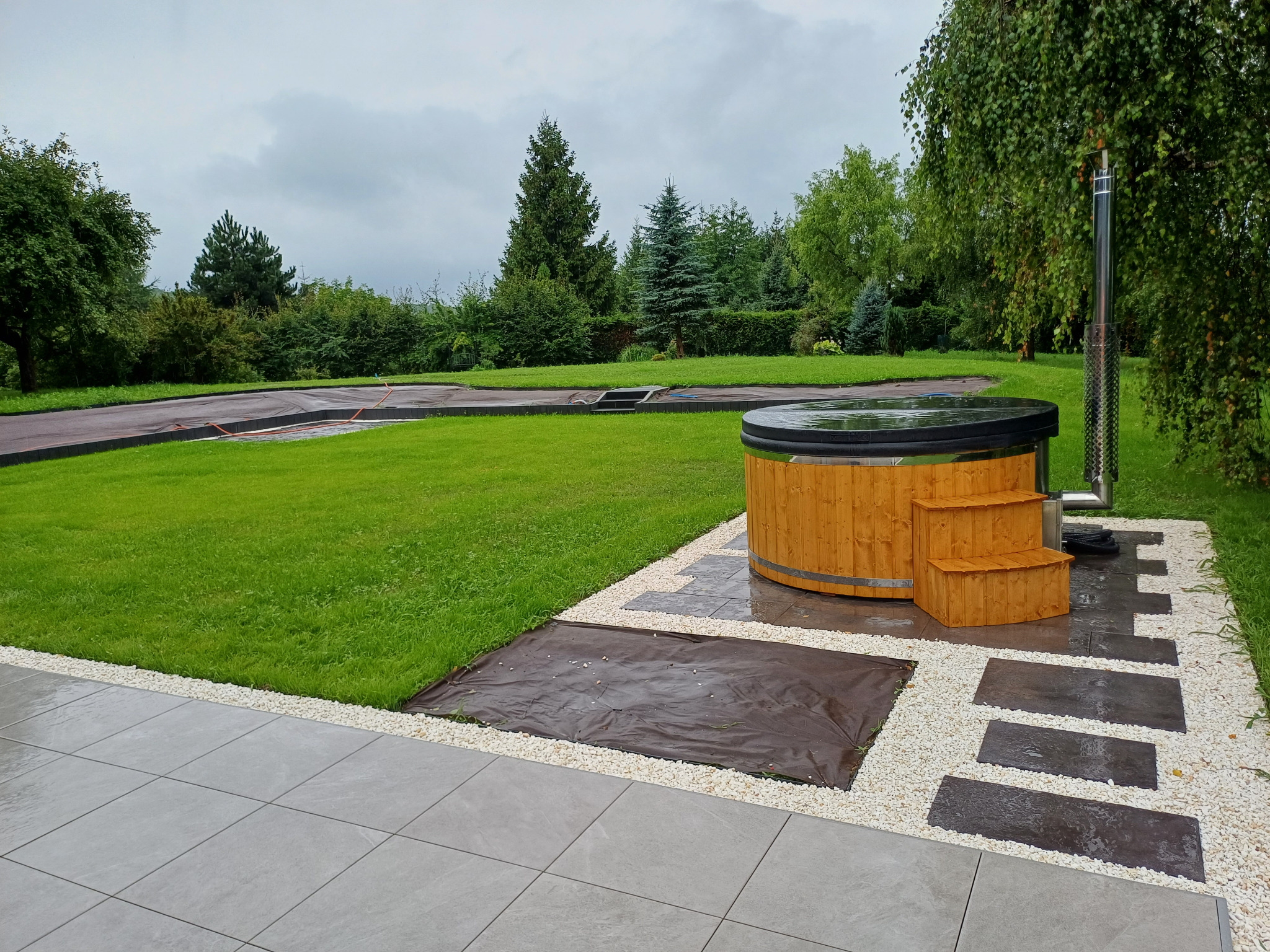 Drewniana balia ogrodowa z metalowym kominem, umieszczona na trawniku z wyłożonymi płytami i białym żwirem, w tle zielony ogród i pochmurne niebo.