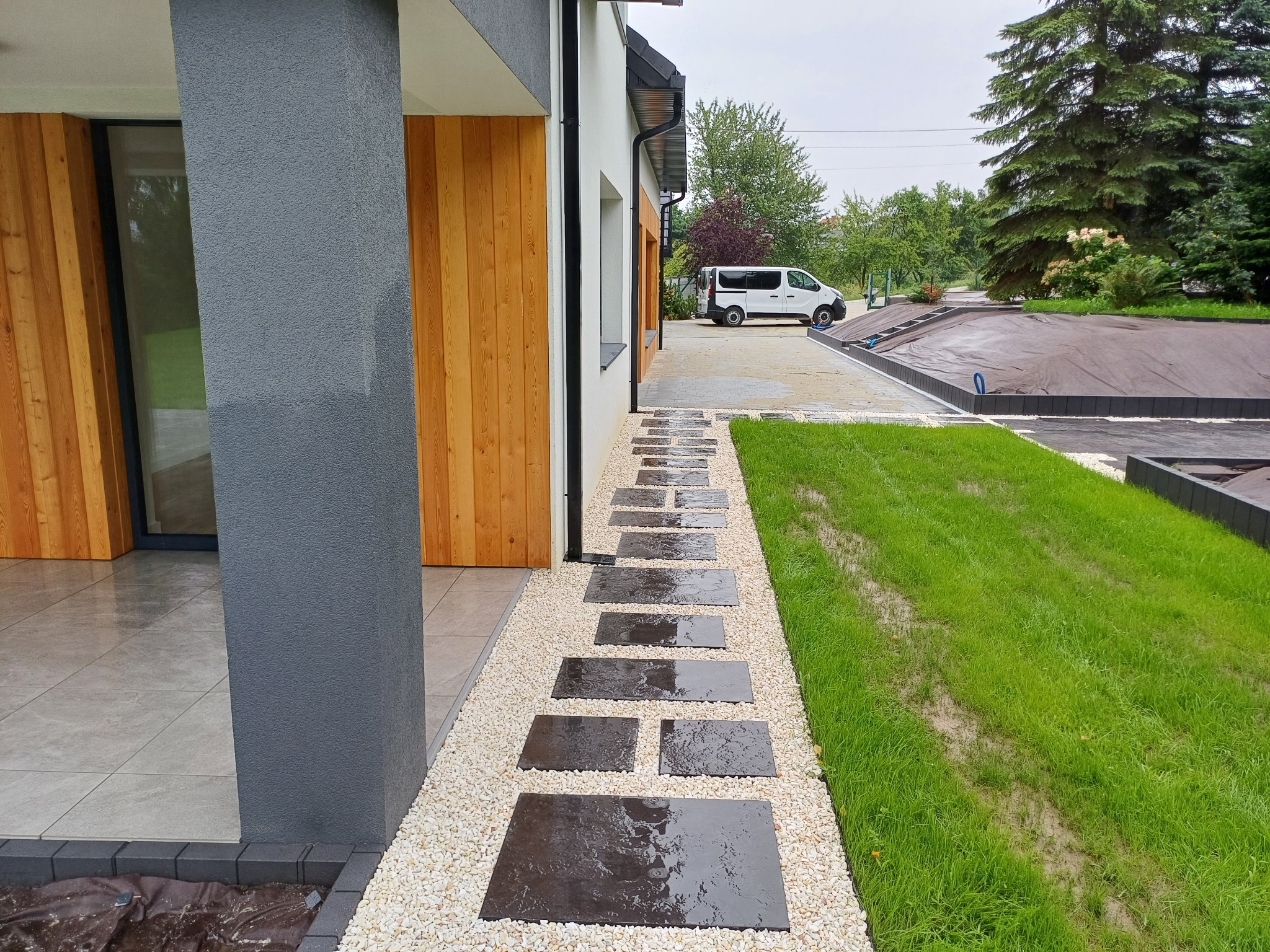 Nowoczesna ścieżka z ciemnych płyt kamiennych ułożona w białym żwirze, prowadząca od budynku z drewnianą elewacją i szarym filarem w kierunku trawnika, z widocznym samochodem dostawczym w tle.