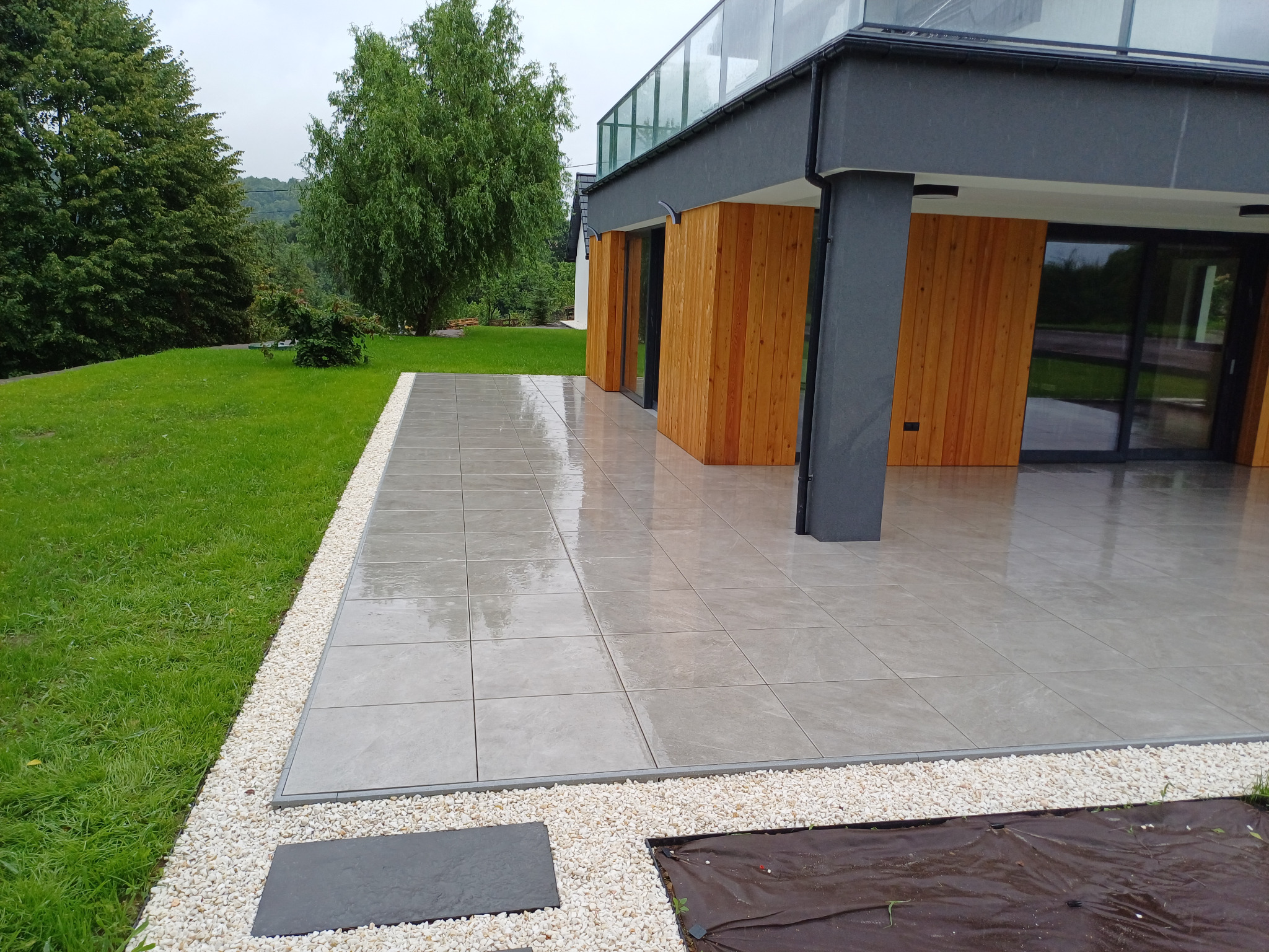 Nowoczesny taras wyłożony szarą, kwadratową kostką brukową, obramowany białym grysem, przylegający do domu z drewnianą elewacją i dużymi oknami.